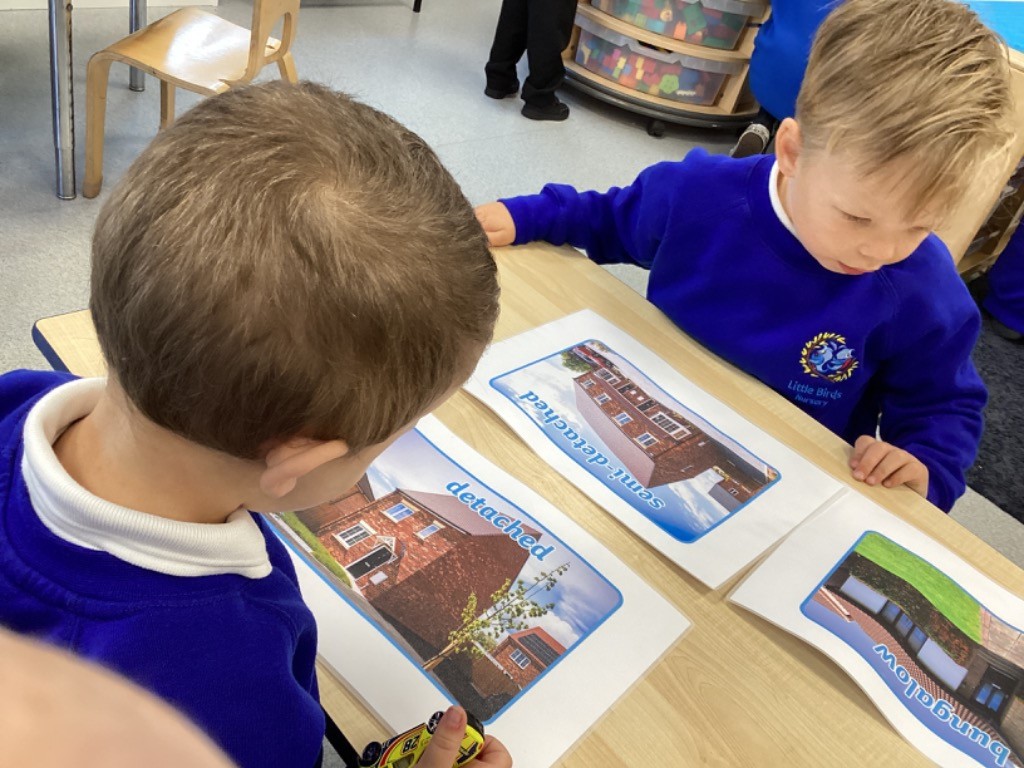 Two children are looking at and discussing similar printouts of houses, labeled "detached," "semi-detached," and "bungalow," furthering the lesson on different dwelling types.