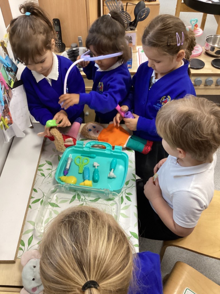 a group of children is absorbed in doctor-themed role-play. They are using various items from the toy doctor's kit, including a stethoscope, to examine a doll and stuffed toys on the table.