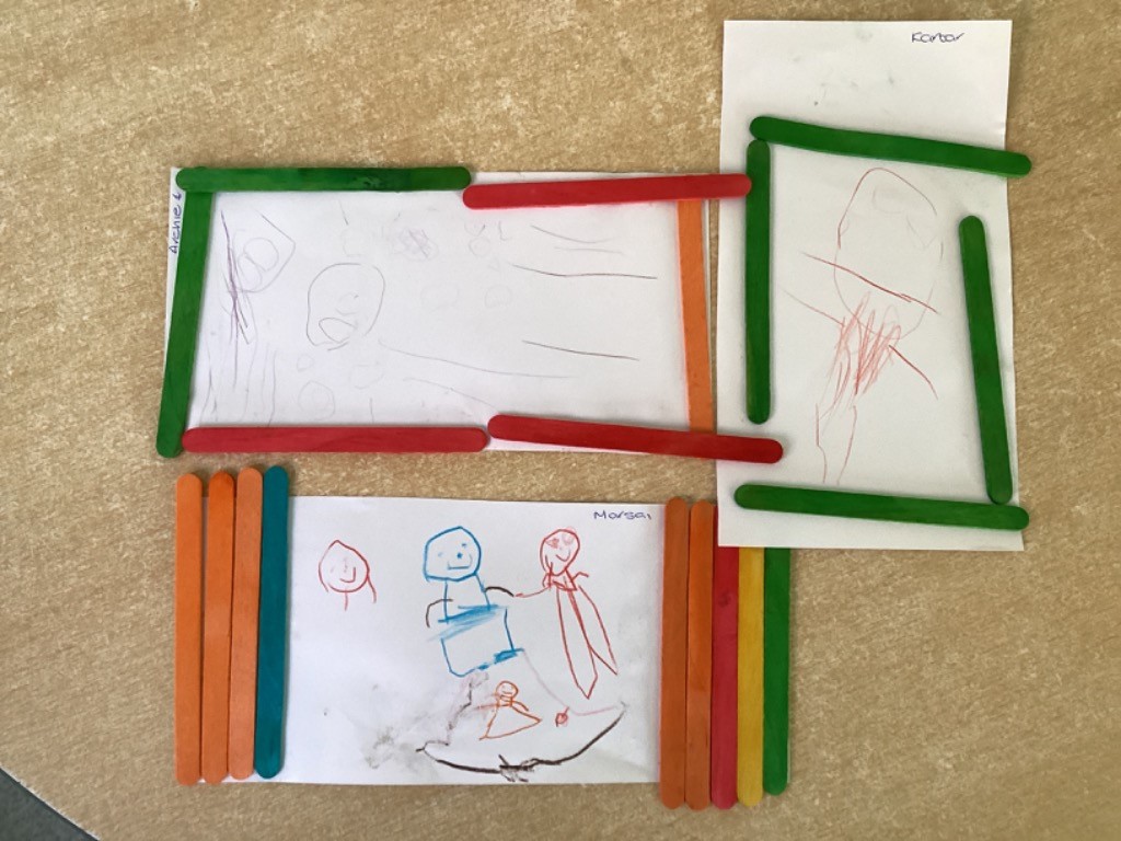 This image captures three finished or in-progress art projects. Children have used brightly colored craft sticks (green, red, orange, blue) to create frames or structures around their own drawings.