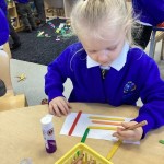 A child is concentrating on arranging and sticking various colored craft sticks (red, yellow, green) onto a piece of white paper. A glue stick is visible nearby.