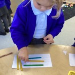 A child is carefully placing a yellow craft stick onto their paper, arranging it alongside a blue and a green stick that are already positioned.