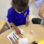 A child is working at a round table, arranging and gluing various colored sticks (green, orange, red, purple) onto paper to create a design.