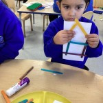 A child is holding up a piece of paper to show their completed construction, which uses sticks to form a shape resembling a simple house or geometric structure with a roof.