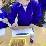 A child is carefully positioning an orange craft stick onto their paper while another child holds a glue stick, suggesting a collaborative or shared activity.