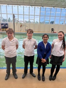 Four children in school uniforms stand side-by-side in front of an indoor swimming pool.