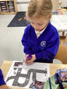 A young child in a blue uniform working on a paper with a road map design, holding a purple object.