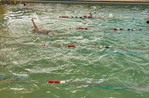 People swimming laps in an indoor pool with lane ropes, with one person in the foreground swimming backstroke and creating a splash.