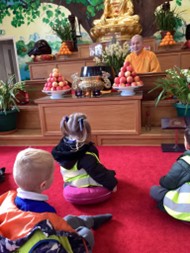 Children sitting on a red carpet facing a monk and a large golden Buddha statue in a temple.