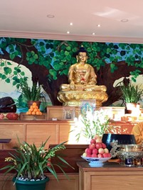 Interior view of a temple with a large golden Buddha statue seated above wooden platforms decorated with flowers and fruit.