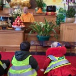 Children in safety vests sitting on the floor in front of a monk in an orange robe inside a temple or shrine.