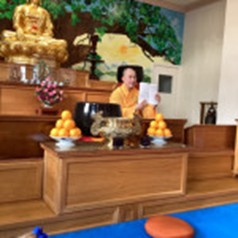 A monk in an orange robe seated on a raised platform holding a paper, with a golden Buddha statue and offerings visible.