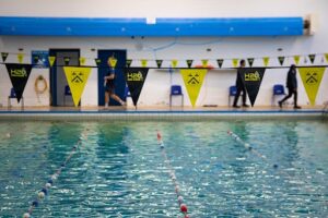 An indoor swimming pool with lane ropes in the foreground. In the background, people walk along the poolside, which is decorated with black and yellow bunting.