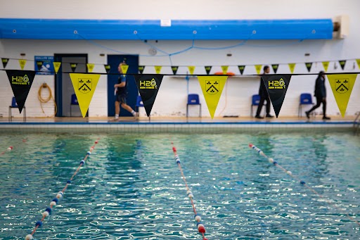 An indoor swimming pool with lane ropes in the foreground. In the background, people walk along the poolside, which is decorated with black and yellow bunting.