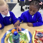 A young girl sitting at a table, holding a small carrot. Trays in front of her contain fruit, a drink, and packaged snacks.