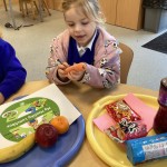 Two young girls in blue tops are reaching towards a tray that holds fruit (banana, orange, apple), a carrot, and a bottle of water. Packaged snacks are on a separate tray.