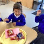 A young girl in a blue uniform sitting at a table, smiling while looking at a tray with packaged snacks and a note or card.