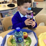 A young boy in a blue uniform sitting at a table, holding and looking at a bottle of drink. A tray in front of him holds a banana, an apple, and another bottle.
