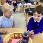 Two young boys in blue uniforms sitting at a table, smiling and holding small food items above a tray of snacks and a drink.