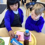 Two young children in blue uniforms looking down at a tray containing snacks and a bottled drink.
