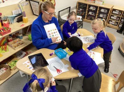 A woman who looks to be a teacher sits a table with 4 children around showing them pictures on a page