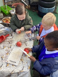Three boys outside in coats cutting up tomatoes on a chopping board and plastic knife