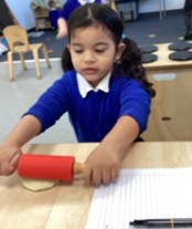 A young girl in a blue uniform with her hair in pigtails, concentrating on rolling out dough with a red miniature rolling pin.