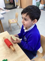 A young boy in a blue uniform sitting at a table, holding a small piece of dough with a miniature red rolling pin next to him.