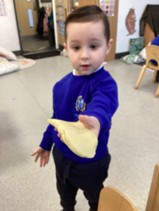 A young boy in a blue uniform standing and holding out a piece of light-colored dough towards the camera.