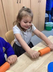 A young girl with light hair, wearing a white polo shirt, smiling as she rolls out dough using a small orange rolling pin on a wooden table.