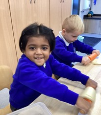 Two young children in blue uniforms smiling while using small rolling pins to work with dough on a wooden table.