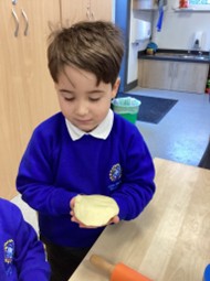 A young boy in a blue uniform holding and looking down at a piece of dough in his hands. An orange rolling pin is visible on the table.
