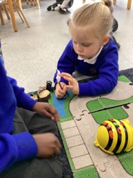 A close-up of a young child lying on the floor, playing with a small toy figure and a yellow robot toy (possibly a Bee-Bot) on a road-themed mat, with another child partially visible on the left.