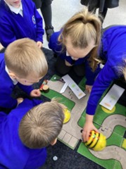 An overhead view of four children gathered on the floor, interacting with a yellow Bee-Bot toy and small cards on a road-themed playmat.