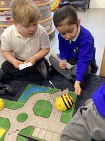 Two children sitting on the floor, holding small cards and looking at a yellow Bee-Bot toy on a road-themed playmat.