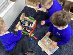 Three young children sitting on the floor, engaging with a nativity scene made from wooden blocks and toys, with one child holding an open book.