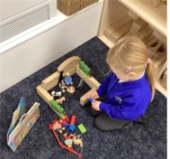 A young child is kneeling on a dark blue carpet, playing with wooden blocks and small animal and figure toys, creating a structure next to an open book.
