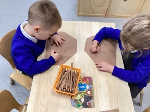 Two young children in blue jumpers sit opposite each other at a table, drawing on brown paper with brown crayons from an orange basket.
