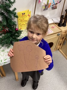 A young child stands next to a small tinsel-decorated Christmas tree, holding up a brown piece of paper with three simple figure drawings.