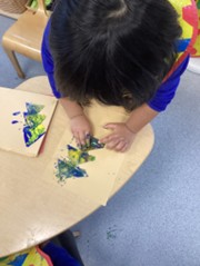 Overhead view of a child using their fingers to smear blue and yellow paint on a piece of light-brown paper at a table.