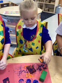 A young child wearing a yellow art smock concentrates on printing with an orange block onto red paper at a table.