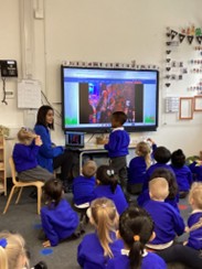An adult teacher and a young student stand by a large interactive screen in a classroom, presenting a photo to a group of children sitting on the floor in blue uniforms.