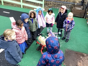 A group of young children in various winter hats and coats stand in a circle outdoors, focused on toys in the center of their grou