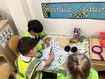 Three children in yellow safety vests sit at a play table marked "Police Station," busy drawing on "WANTED" posters.