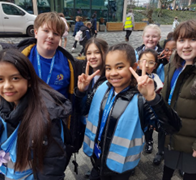 A group of several school-aged children posing outdoors; many are smiling and making peace signs with their hands while wearing blue lanyards and vests.