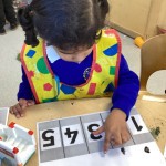 A child wearing a colorful shape-patterned apron works on a counting activity involving numbers 1 through 5.