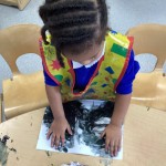 A child with braided hair and a play apron uses their hands to create a messy black paint or ink artwork on paper.