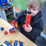 A child in a black coat constructs a tower using red, blue, and yellow plastic blocks at an outdoor table.