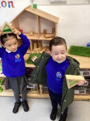Two children in school uniforms hold triangular blocks while standing in front of a wooden dollhouse.