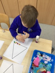 A child in a school uniform uses a glue stick to attach small, colorful decorations to a large triangle printed on paper.