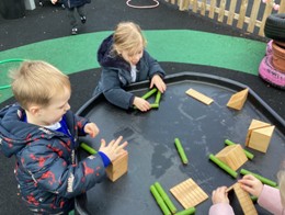 Three children play with wooden blocks and green cylinders on a black outdoor activity table.